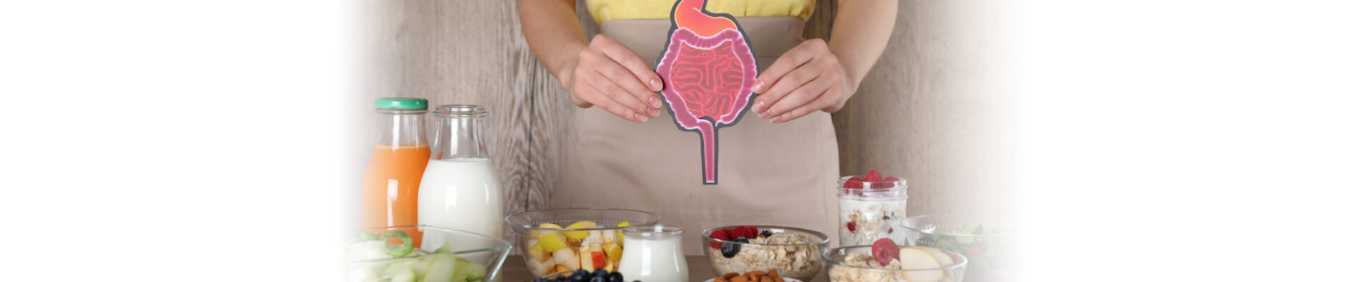 Una persona vestida con un delantal claro sostiene un dibujo del sistema digestivo sobre su abdomen. Delante de ella, sobre una mesa de madera, hay varios cuencos y vasos con distintos alimentos diuréticos y saludables, como frutas (arándanos, frambuesas), cereales, leche, zumo de naranja, ensalada y frutos secos, listos para ser consumidos.