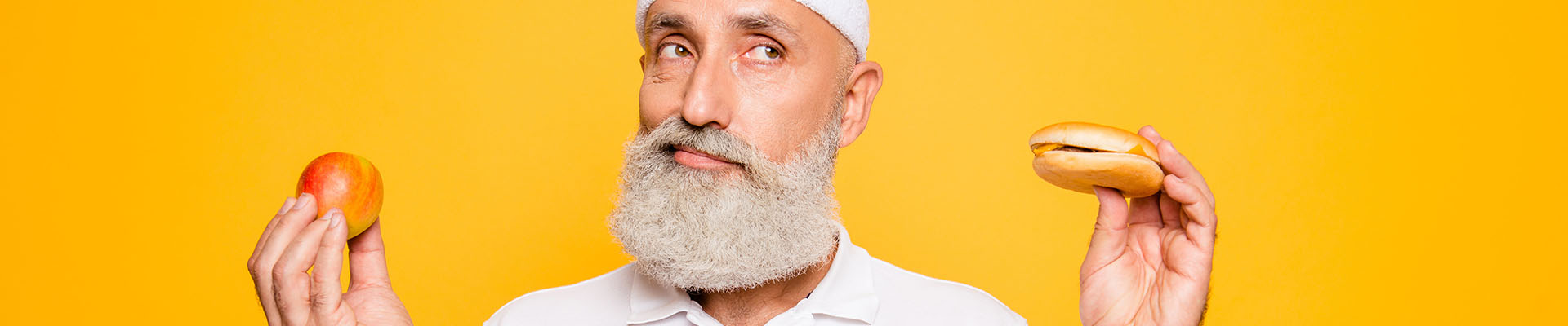 Imagen de un hombre mayor vestido de blanco en una habitación de color naranga. Tiene una hamburguesa en una mano y una manzana en la otra y quiere representar la diferencia marcada entre alimentos prohibidos para la incontinencia urinaria y los recomendables.