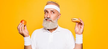 Imagen de un hombre mayor vestido de blanco en una habitación de color naranga. Tiene una hamburguesa en una mano y una manzana en la otra y quiere representar la diferencia marcada entre alimentos prohibidos para la incontinencia urinaria y los recomendables.