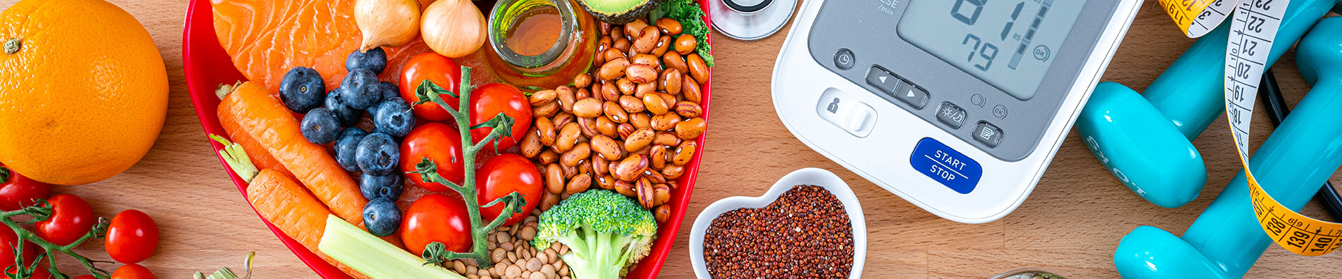 Conjunto de alimentos que bajan el colesterol repartidos por encima de una mesa, algunos están en un plato con forma de corazón. En la mesa también hay objetos vitales para el colesterol, por ejemplo un estetoscopio, un tensiómetro y unas pesas, que quieren simbolizar el ejercicio.
