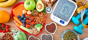 Conjunto de alimentos que bajan el colesterol repartidos por encima de una mesa, algunos están en un plato con forma de corazón. En la mesa también hay objetos vitales para el colesterol, por ejemplo un estetoscopio, un tensiómetro y unas pesas, que quieren simbolizar el ejercicio.