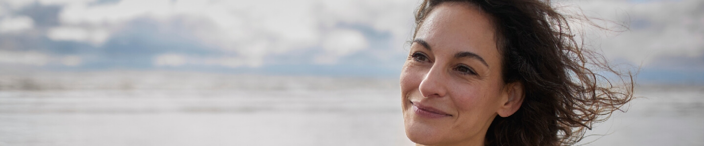 Mujer en una playa mirando felizmente al horizonte, ya que se siente inspirada. Ha recibido apoyo psicológico y ahora ve el mundo de una forma más optimista.