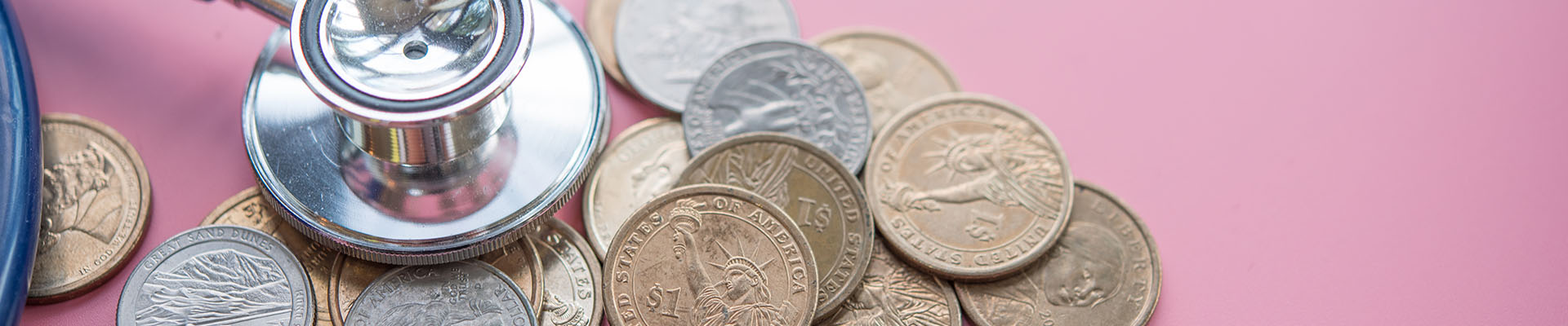 Estetoscopio apoyado en unas monedas que están esparcidas por una mesa rosa. Esta imagen quiere ser una metáfora de los beneficios fiscales que tienen algunos enfermos como los discapacitados.