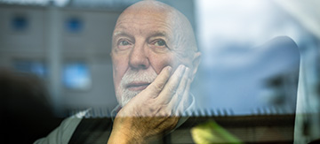 Fotografía a través de una ventana, al otro lado en el interior de un hogar un hombre mayor con calvicie mira con tristeza el exterior. Tiene incontinencia urinaria por distintas causas y esto le genera problemas en su vida.