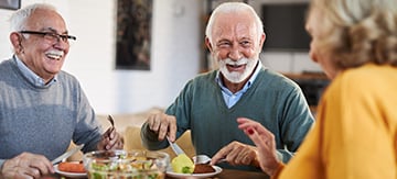 Dos hombres mayores y una mujer mayor compartiendo una comida juntos en una mesa. Están pasando un buen rato juntos, al mismo tiempo que comen alimentos saludables buenos para aumentar el colesterol bueno HDL.