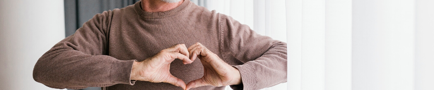 Hombre mayor haciendo con sus manos el gesto de corazón, justo encima de su pecho donde se encuentra este órgano vital. Lo hace para mostrar que está luchando con su colesterol y actualmente se encuentra mejor ya que tiene niveles de colesterol saludables para su edad.