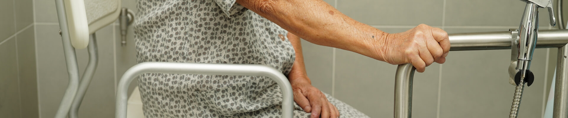 Imagen de cómo lavar una persona con movilidad reducida. Una mujer mayor se encuentra en el baño sentada en su silla de ducha y con la mano sostenida en la barandilla para tener más estabilidad de personas con movilidad reducida.