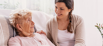 Chica joven al lado de la cama donde descansa su madre, de la tercera edad. La chica es de gran ayuda para esta, ya que le ayuda en su día a día, sabe como poner un pañal a un adulto entre otras cosas. Es por esto que están contentas.