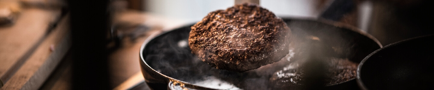 Hombre cocinando una hamburguesa en una paella, en el momento de la imagen justo la está girando. Sabe prepararla gracias a consejos y recetas que ha encontrado en una página por internet.