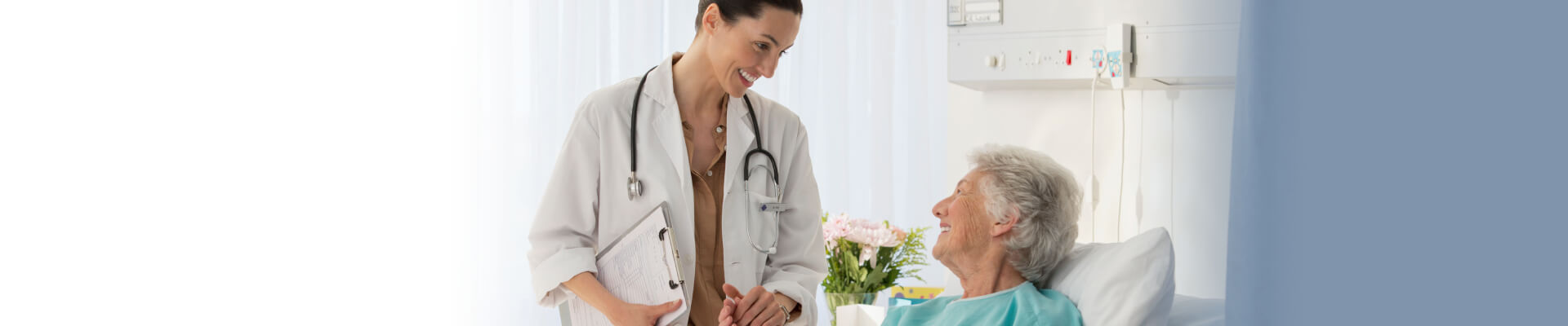 Mujer tumbada en su cama del hospital contenta y mirando a su doctora, que está justo de pie al lado izquierdo de su cama sosteniendo su mano y sonriendo a la paciente. Las dos están contentas ya que la operación ha sido un éxito y la doctora ha contestado a la pregunta de cuánto tiempo dura la incontinencia después de una operación de próstata. 