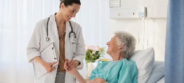 Mujer tumbada en su cama del hospital contenta y mirando a su doctora, que está justo de pie al lado izquierdo de su cama sosteniendo su mano y sonriendo a la paciente. Las dos están contentas ya que la operación ha sido un éxito y la doctora ha contestado a la pregunta de cuánto tiempo dura la incontinencia después de una operación de próstata.