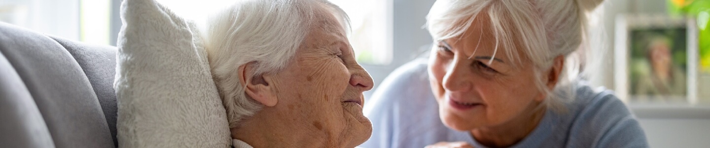 Mujer de unos 60 años sonriendo a su madre de unos 90 años que se encuentra sentada en un sofá. La madre es dependiente de la hija, pero aún así tienen una familia feliz ya que se quieren.