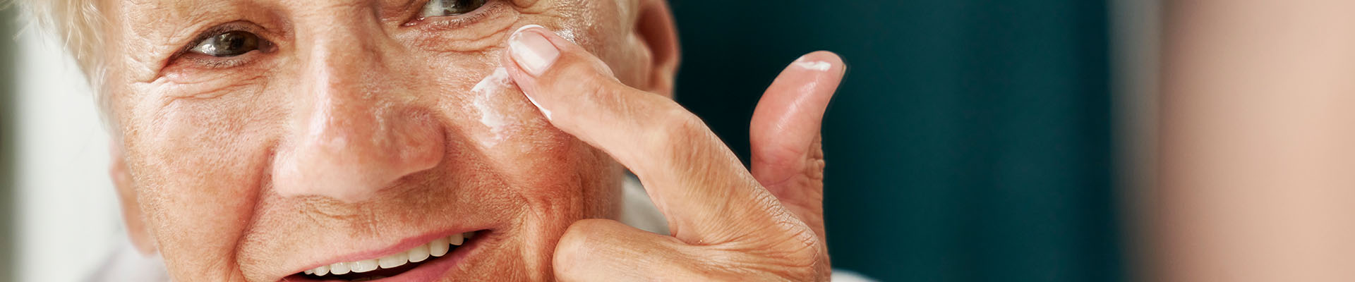 Mujer mayor aplicando una capa de crema en su mejilla. La crema es importante para el cuidado de la piel, especialmente si sufrimos algún tipo de roce, como podria ser la dermatitis del pañal en adultos por ejemplo.