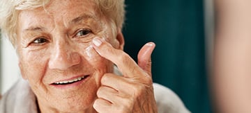 Mujer mayor aplicando una capa de crema en su mejilla. La crema es importante para el cuidado de la piel, especialmente si sufrimos algún tipo de roce, como podria ser la dermatitis del pañal en adultos por ejemplo.