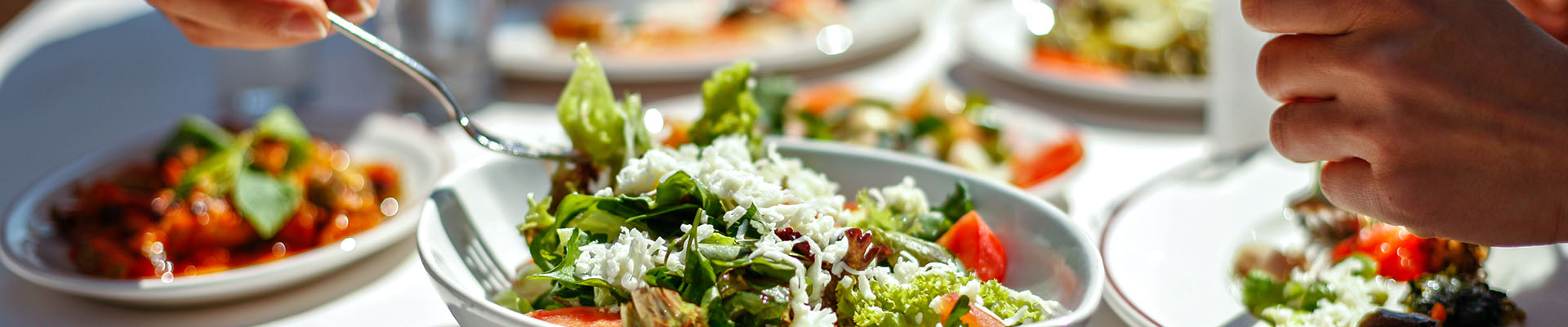 Mesa de restaurante llena de platos sabrosos y variados, con mucho verde y las manos de dos personas que comen estos platos. Todos estos platos son saludables y podrían formar parte de una dieta para reducir el colesterol.