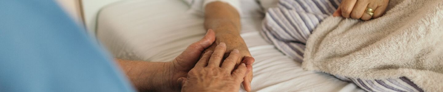 Hombre mayor sentado al lado de una cama, en la que se encuentra su mujer, que es una paciente encamada. Se dan la mano cariñosamente ya que, se quieren a pesar de la enfermedad.