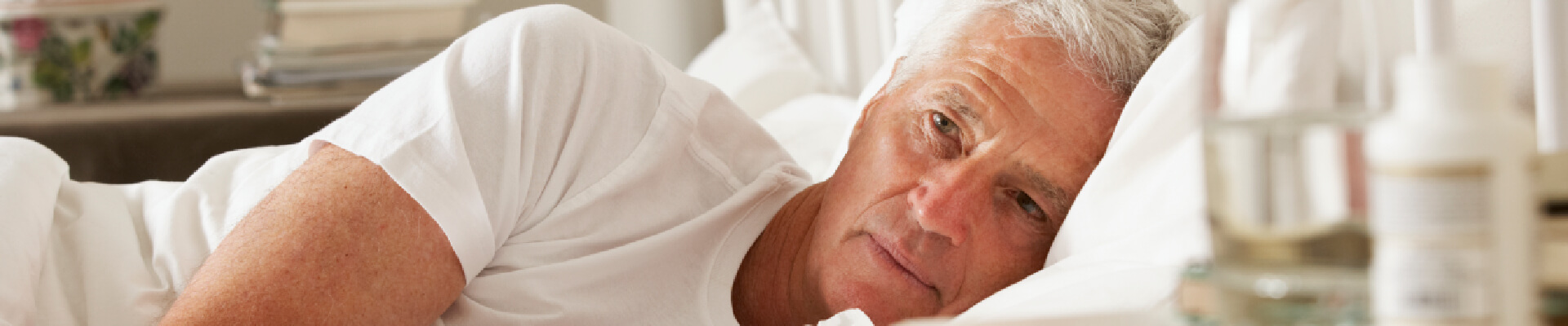 Hombre tumbado en su cama con cara de preocupado. Su cama de sábanas blancas y su pijama blanco también, han sido manchados y se acaba de dar cuenta. Sufre de enuresis nocturna y es por esto que ha cogido un pañuelo, para intentar limpiar un poco lo marcado por su problema.