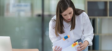 Chica joven vestida de camisa hurgando entre un montón de papeles. Su cara muestra preocupación ya que es difícil conseguir la excedencia por cuidado de familiares.