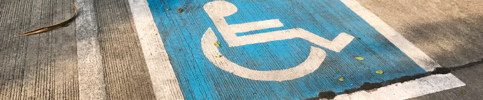Esta imagen muestra en primer plano una plaza de parking en plena calle destinada para personas con algún tipo de discapacidad, por ejemplo incapacidad permanente por incontinencia fecal. Sabemos que es una plaza de parking para minusválidos, ya que en medio de las líneas hay un cuadrado azul con un dibujo esquemático de una persona en silla de ruedas. 