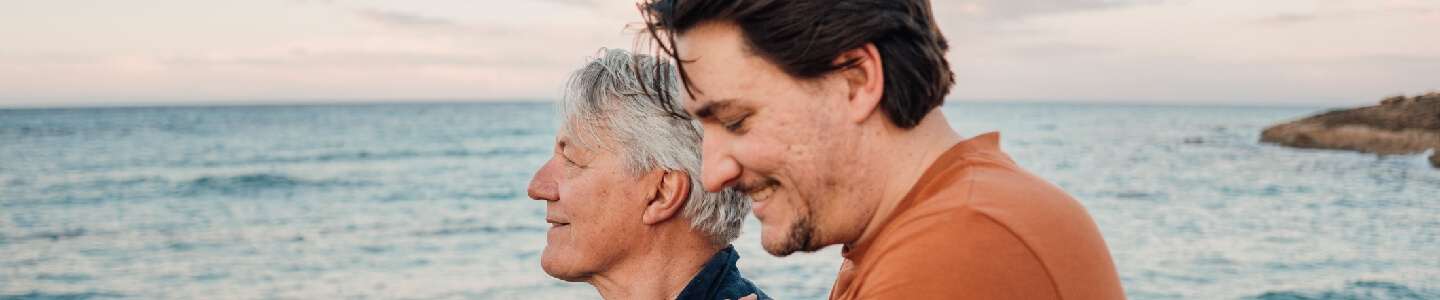 Hijo abrazando a su padre de edad avanzada, el cual sufre de incontinencia ligera, mientras disfrutan de un agradable paseo por una playa de su ciudad.