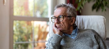 Hombre sentado en el sillón de su casa mirando con cara de preocupación la ventana y apoyando su cabeza en su mano. Está preocupado por el hecho que sufre incontinencia urinaria en hombres. Esto le supone un conjunto de problemas en el dia a dia, pero aún así puede encontrar alguna solución como los ejercicios pensados para solucionar su problema.