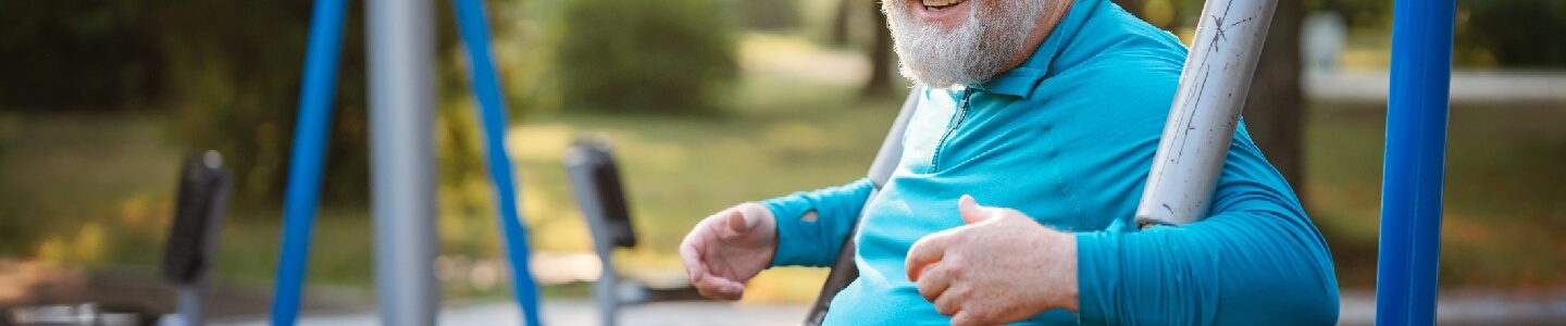 Hombre mayor con obesidad en un parque con máquinas de ejercicio. El señor lleva ropa deportiva y parece estar disfrutando de hacer ejercicio ya que sonríe a la cámara.