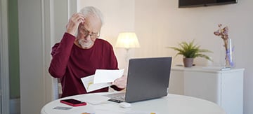 Hombre mayor con un papel en la mano y frente al ordenador, rascándose la cabeza, tratando de entender cómo pasar de incapacidad permanente total a jubilación.
