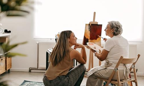 Chica joven acompañando a una mujer mayor en su pasatiempo. Pintar un lienzo blanco y hacer volar la imaginación, las dos lo pasan muy bien pintando.