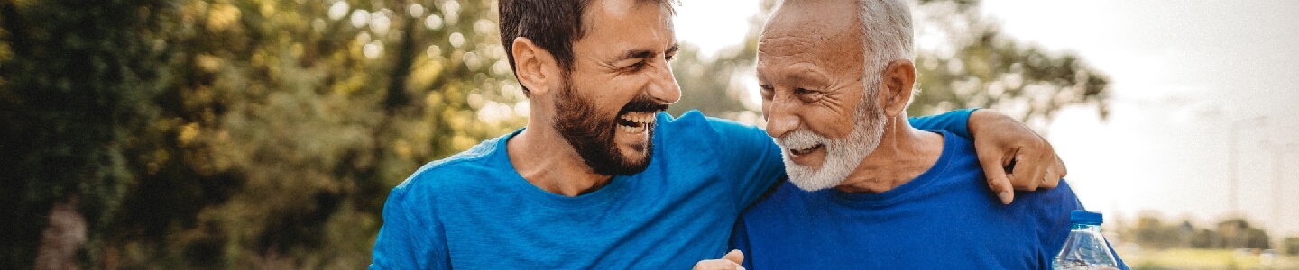 Padre e hijo celebran que han ido a correr, para cuidar su salud cardiovascular. El hijo y el padre sonríen, mientras el hijo abraza por el hombro a su padre. Ambos llevan botellas de agua ya que van a hidratarse después del ejercicio físico.