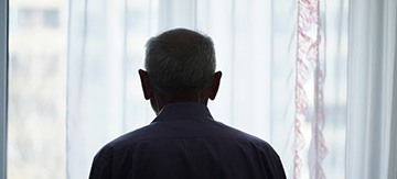 Hombre mayor de espaldas mirando triste por la ventana de la habitación de su casa ya que no quiere ir a una residencia.